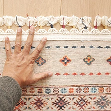 Illustration - Ihr Experte für schönes Wohnen - Eine Nahaufnahme einer Hand, die auf einem wunderschönen, farbenfrohen Vintage-Teppich aus der Vergangenheit platziert ist.